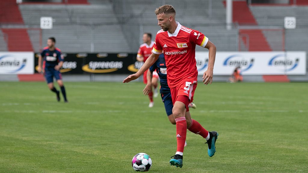 Ein Spieler in roter Fußballuniform mit einer Kapitänsbinde dribbelt den Ball auf einem Rasenfeld während eines Spiels.