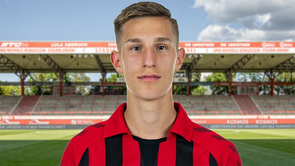 Ein junger Mann mit Kurzhaarschnitt trägt ein rot-schwarz gestreiftes Fußballtrikot vor einem Stadionhintergrund.
