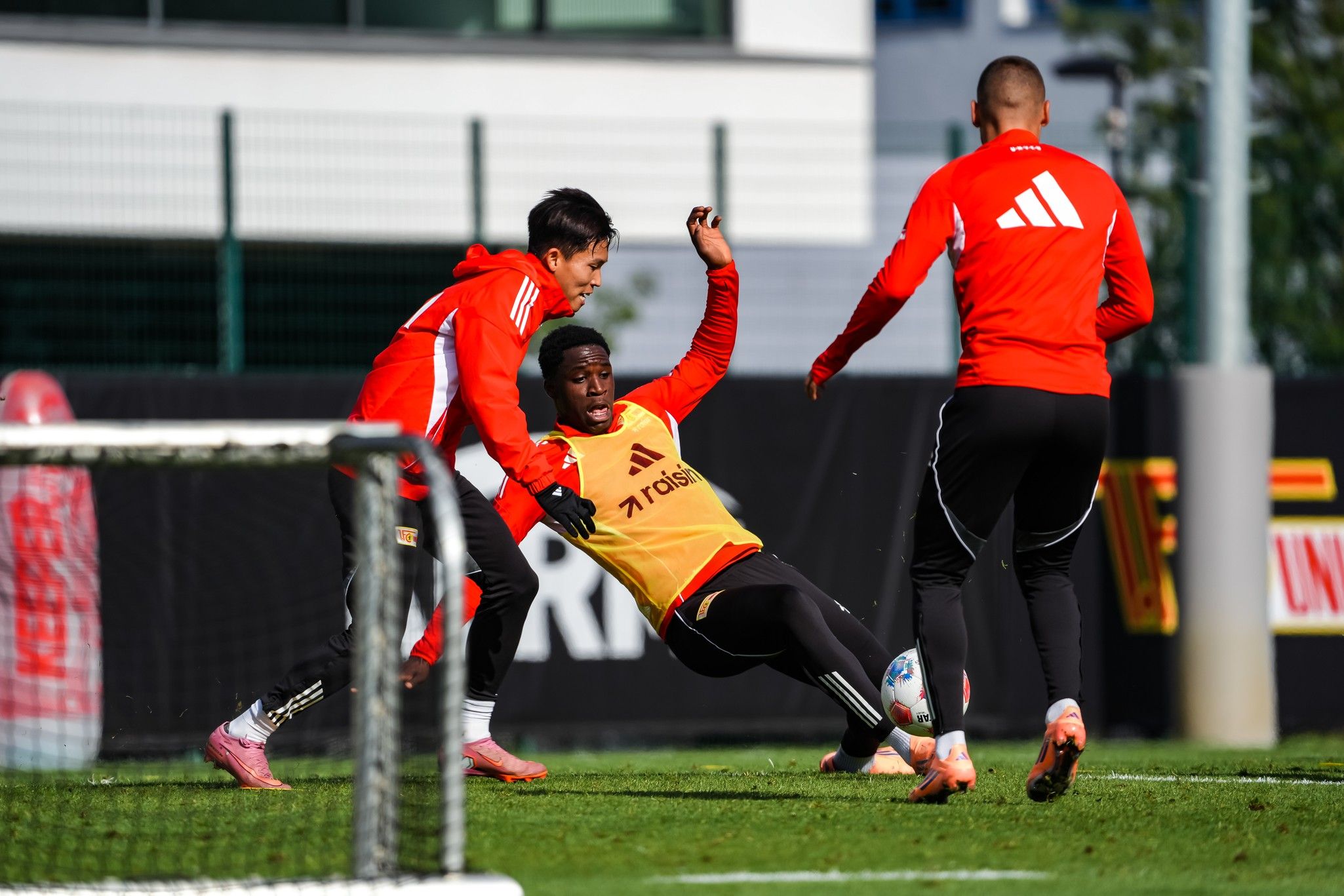 Drei Fußballspieler in Trainingskleidung, einer fällt auf den Boden, während die anderen ihn unterstützen, auf einem Trainingsfeld.