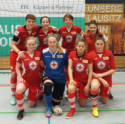 Eine Fußballmannschaft in roten Trikots posiert in einer Sporthalle. Im Vordergrund steht die Torhüterin in blau.