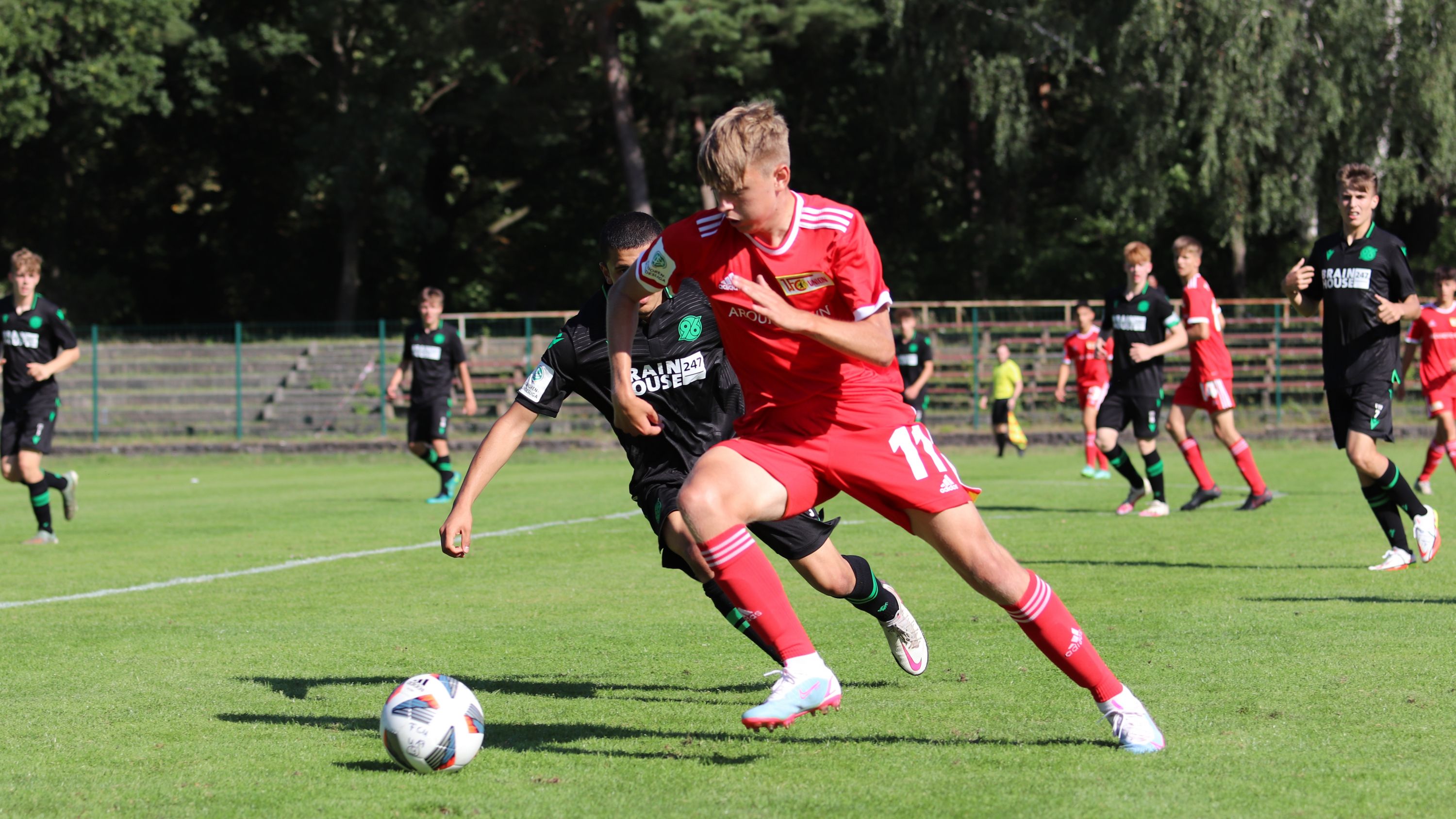 Ein Fußballspiel mit zwei Spielern: Einer in rotem Trikot dribbelt den Ball, während der andere in Schwarz ihn verfolgt.