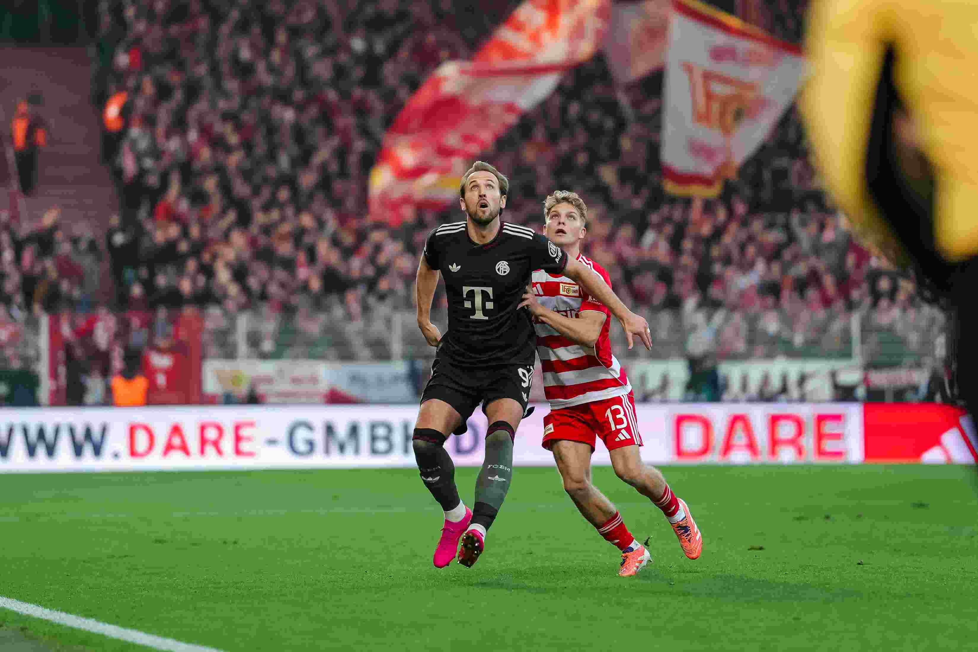 Ein Spieler in Schwarz trägt ein Trikot. Ein Spieler in Rot hält ihn fest. Im Hintergrund sind ein Stadion und Fans.