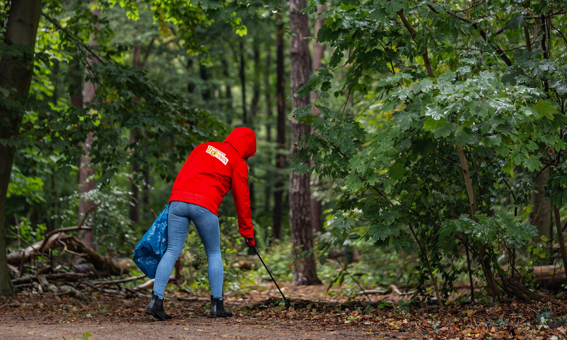 Eine Person in einer roten Jacke sammelt Müll im Wald, während sie einen blauen Sack hält.