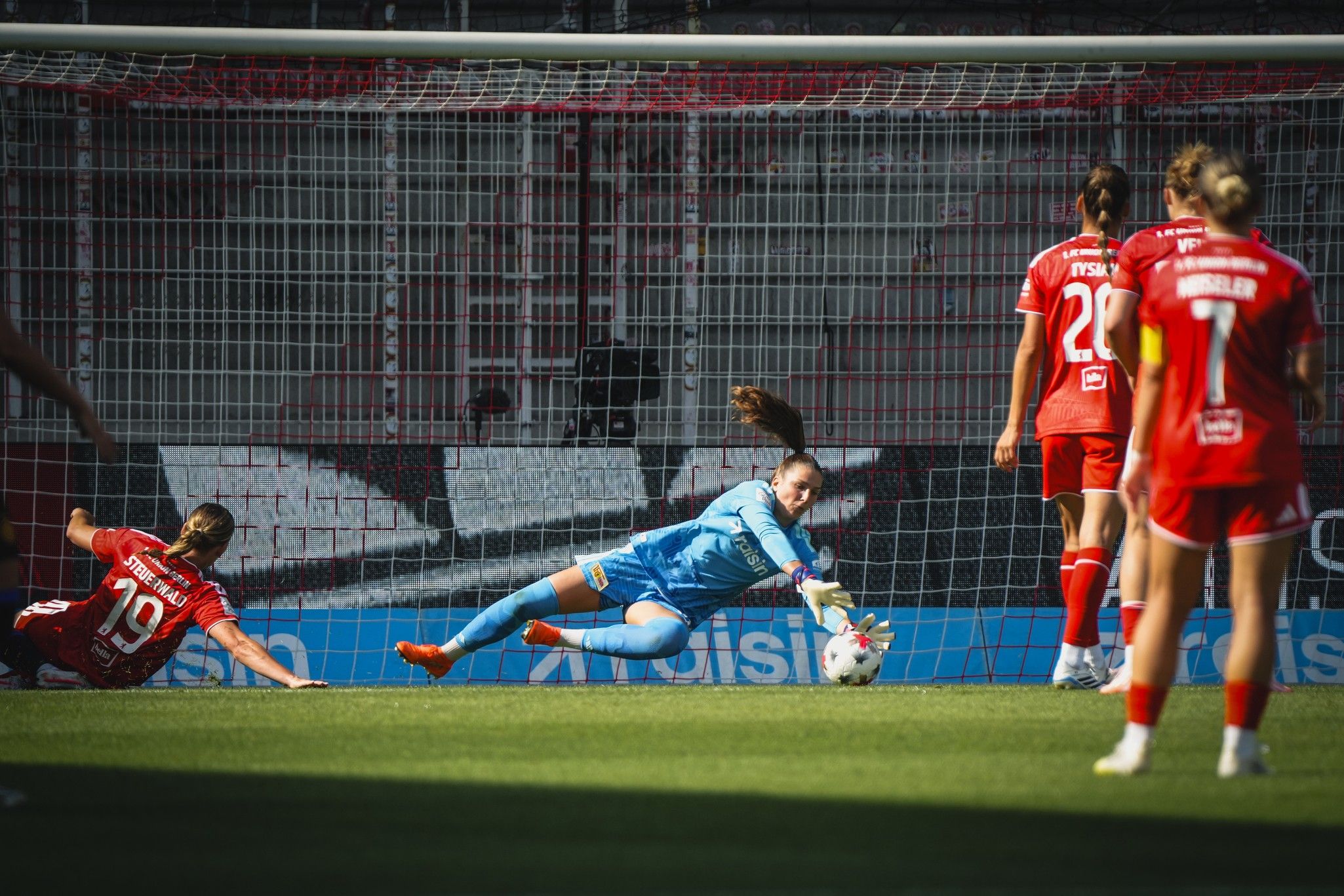 Torhüterin springt, um einen Schuss zu parieren, während Spielerinnen im Hintergrund stehen.