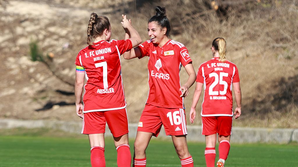 Three female soccer players from 1. FC Union Berlin in red jerseys celebrate a goal on the field.