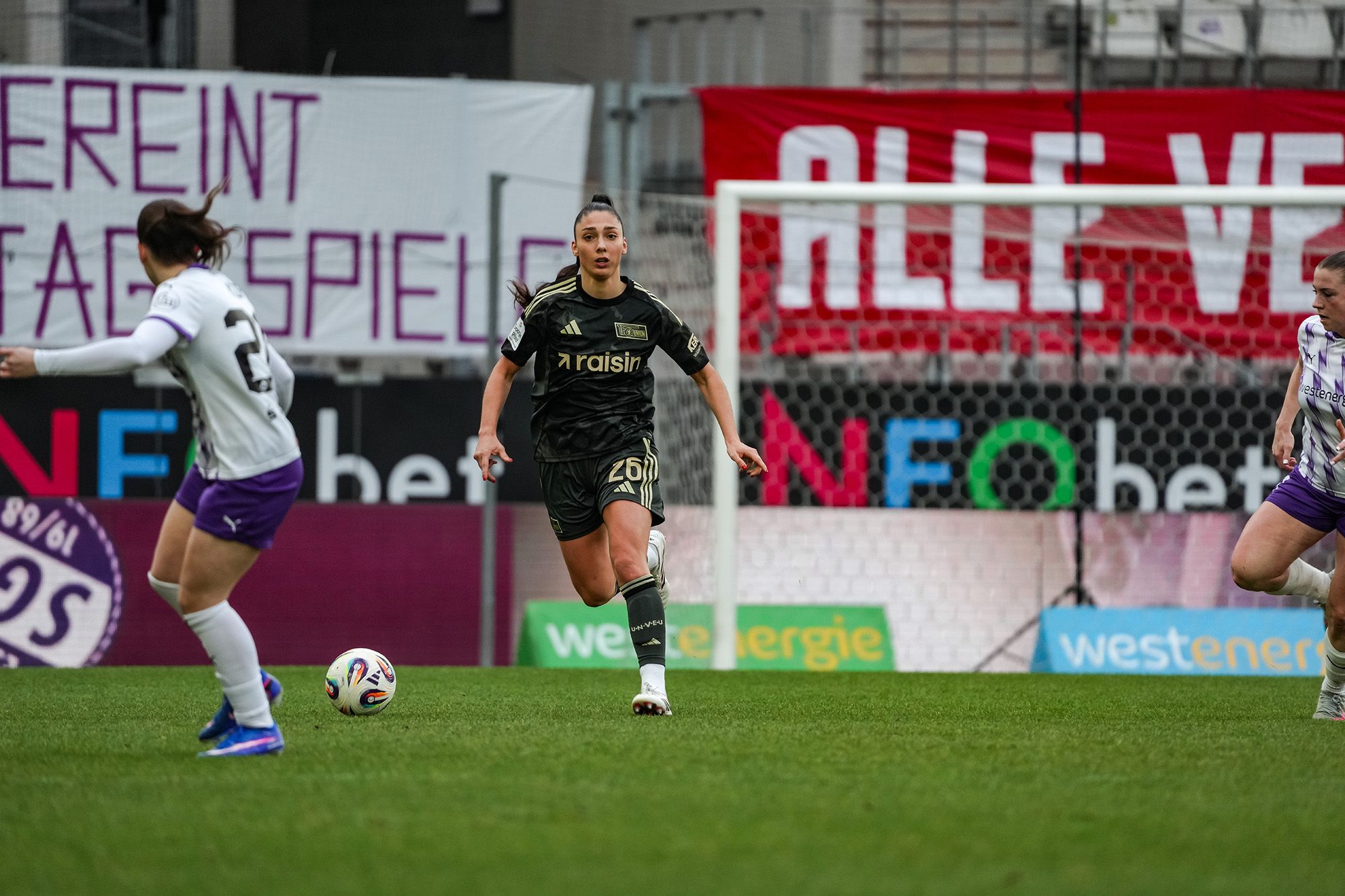 Spielszene im Frauenfußball: Eine Spielerin im schwarzen Trikot dribbelt den Ball, während eine Gegnerin in einem weißen Trikot Nahaufnahme hat.
