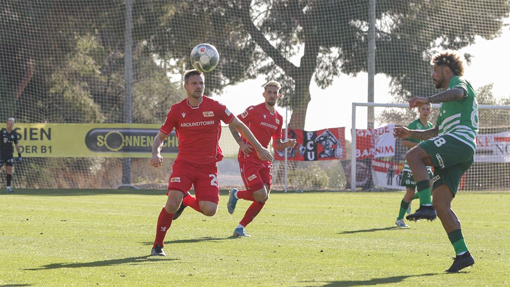 Zwei Fußballspieler in roten Trikots verfolgen einen Ball, während ein Gegner in grünem Trikot zuschießt.
