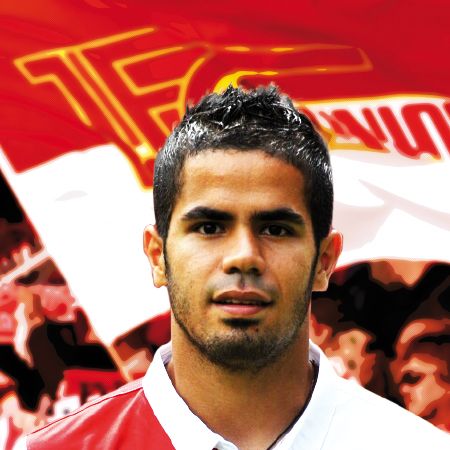 Porträt eines jungen Mannes mit dunklem Haar, vor einem verschwommenen Hintergrund in Rot und Weiß, der ein Fußballtrikot trägt.