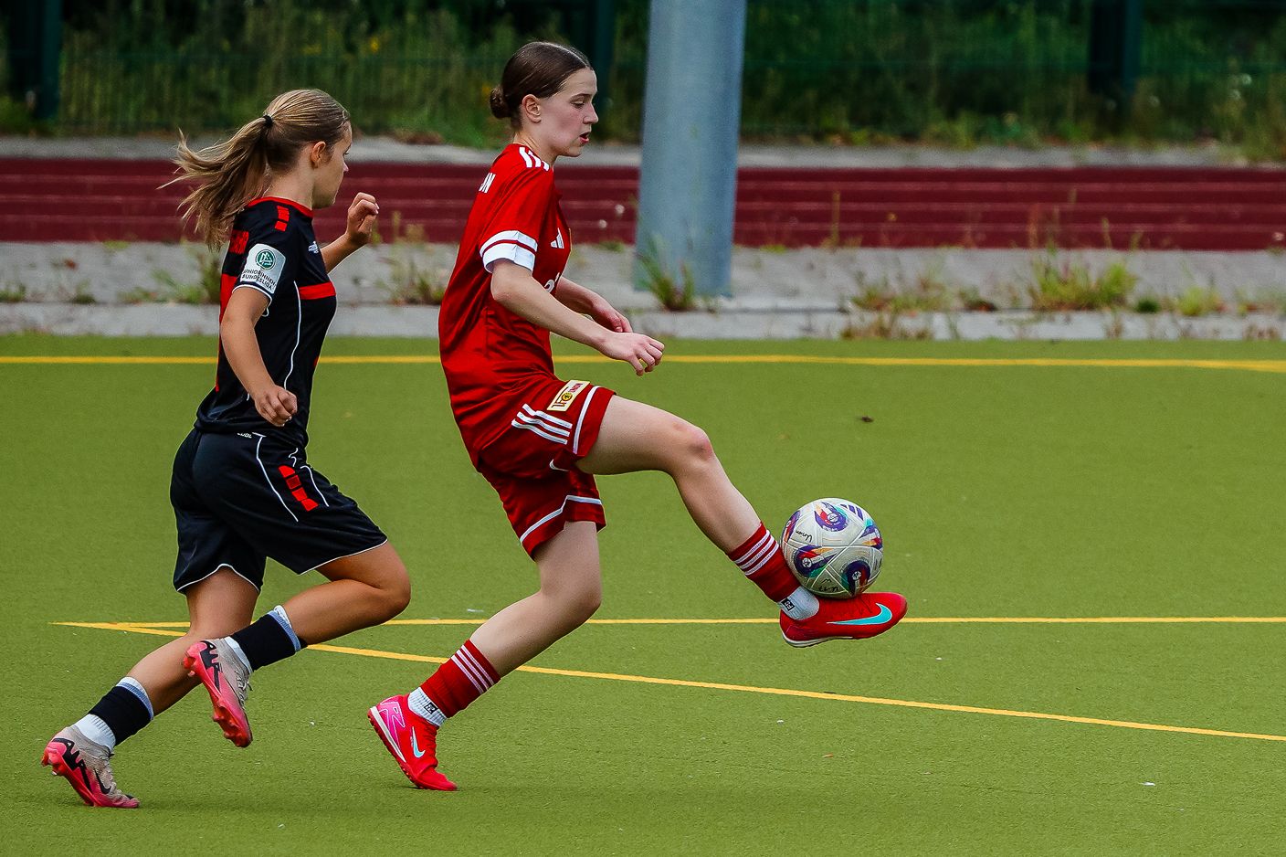 Zwei junge Fußballspielerinnen in Aktion; eine in rotem Outfit dribbelt den Ball, die andere in schwarzem Outfit folgt.