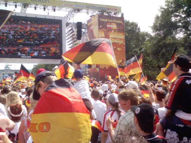 Menschenmenge mit Schwarz-Rot-Gold-Fahnen, die vor einer großen Leinwand bei einem Ereignis in Berlin feiern.