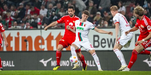 Zwei Fußballspieler im Wettkampf um den Ball während eines Spiels, mit einem weiteren Spieler im Hintergrund.