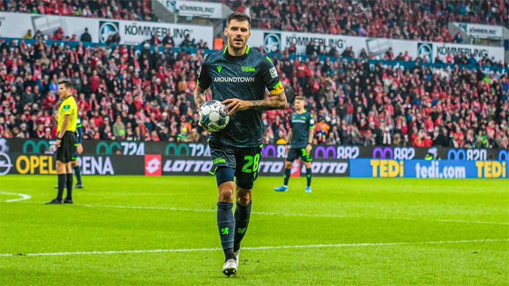 Ein Fußballspieler in dunklem Trikot hält den Ball in der Hand, umgeben von jubelnden Fans im Stadion.