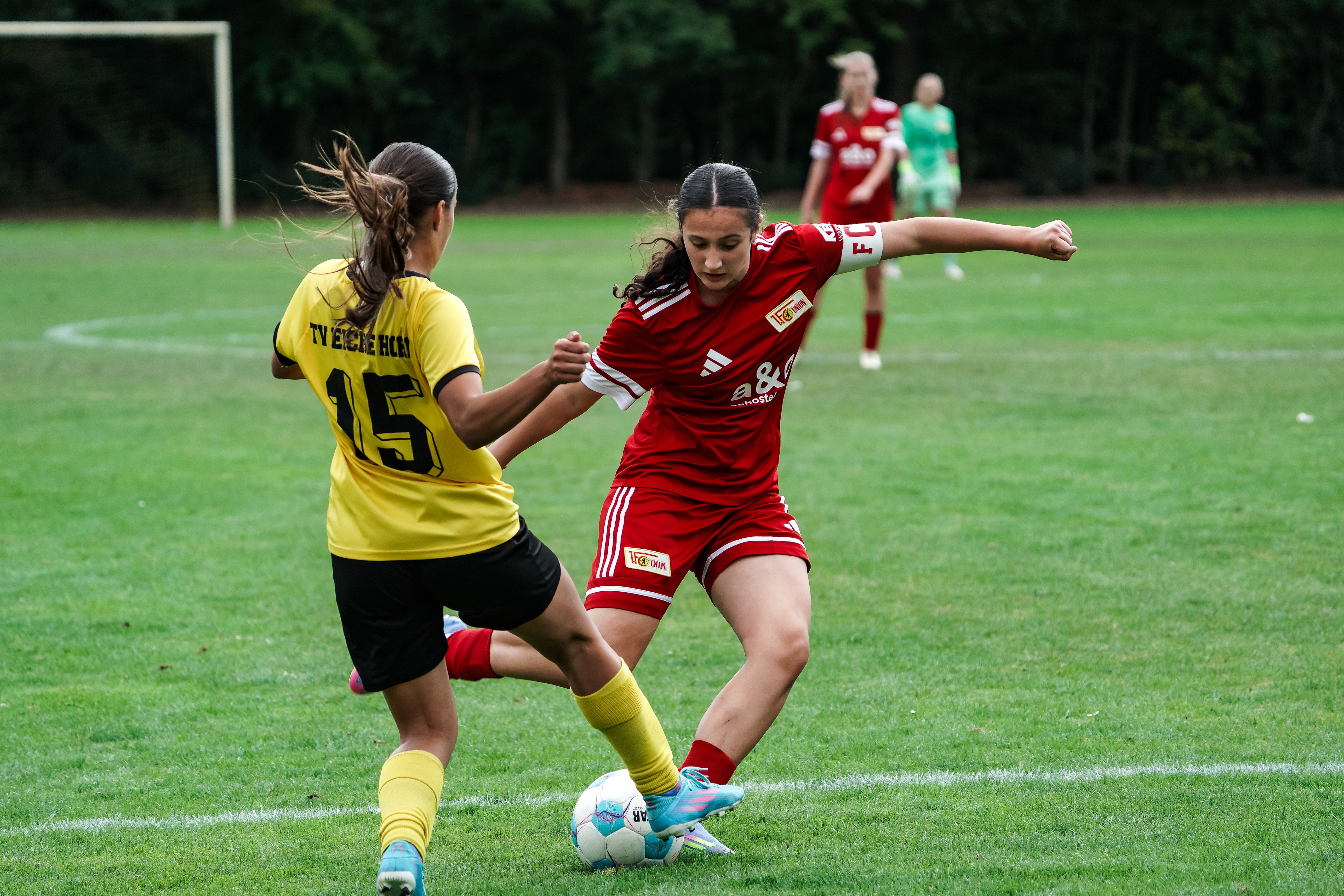 Two female soccer players competing for the ball on a grass field. One player is wearing red, the other yellow.