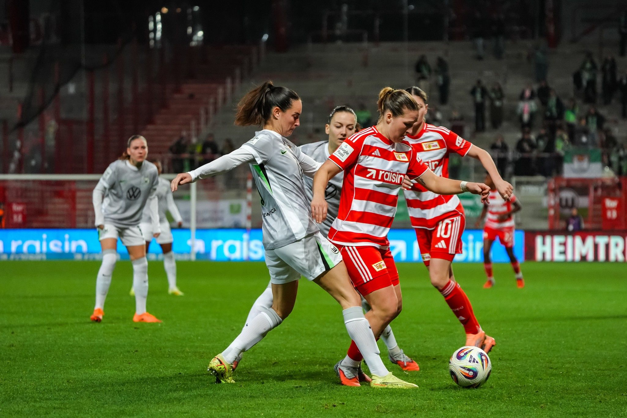 Drei Fußballspielerinnen im Wettkampf um den Ball auf einem Spielfeld, umgeben von Zuschauern.