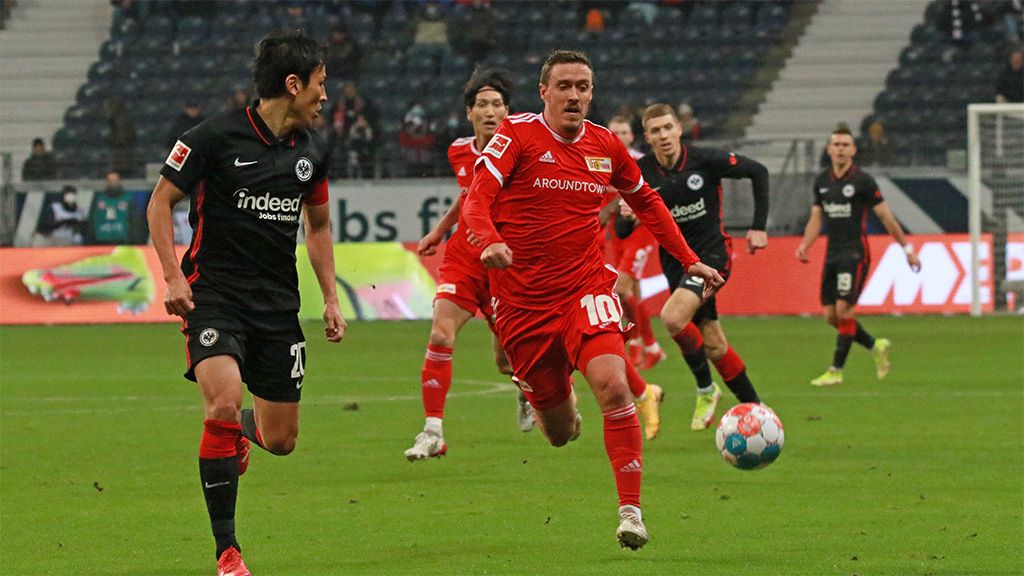 A soccer player in red jerseys dribbles the ball while a player in black chases him. Background with spectators in the stadium.