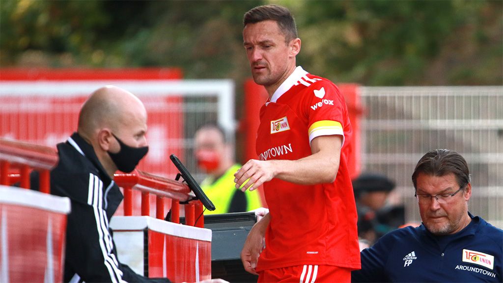 Ein Fußballspieler in rotem Trikot verlässt die Bank, während zwei Betreuer auf ihn achten.