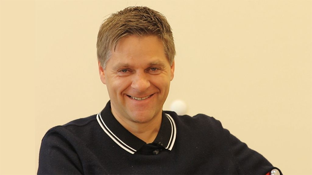 A man with short, light brown hair and a broad smile is wearing a black polo shirt with a white collar.