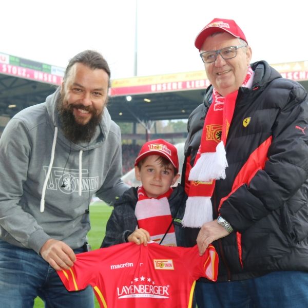 Drei Personen in Fußballkleidung und Schals posieren in einem Stadion, während ein Junge ein Trikot hält.