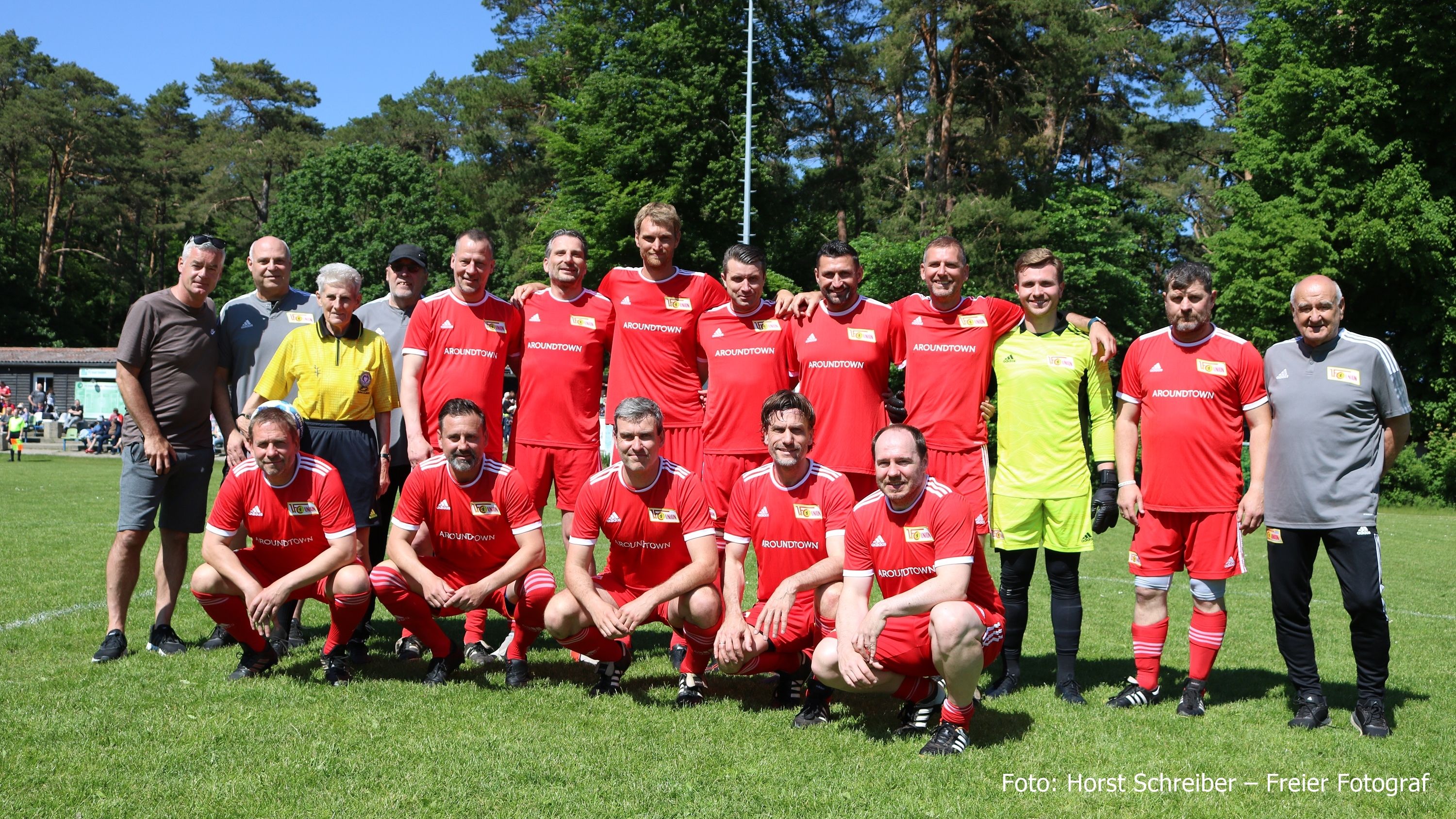 Eine Gruppe von 16 Fußballspielern in roten Trikots posiert auf einem Rasenplatz, umgeben von Bäumen und Zuschauern.