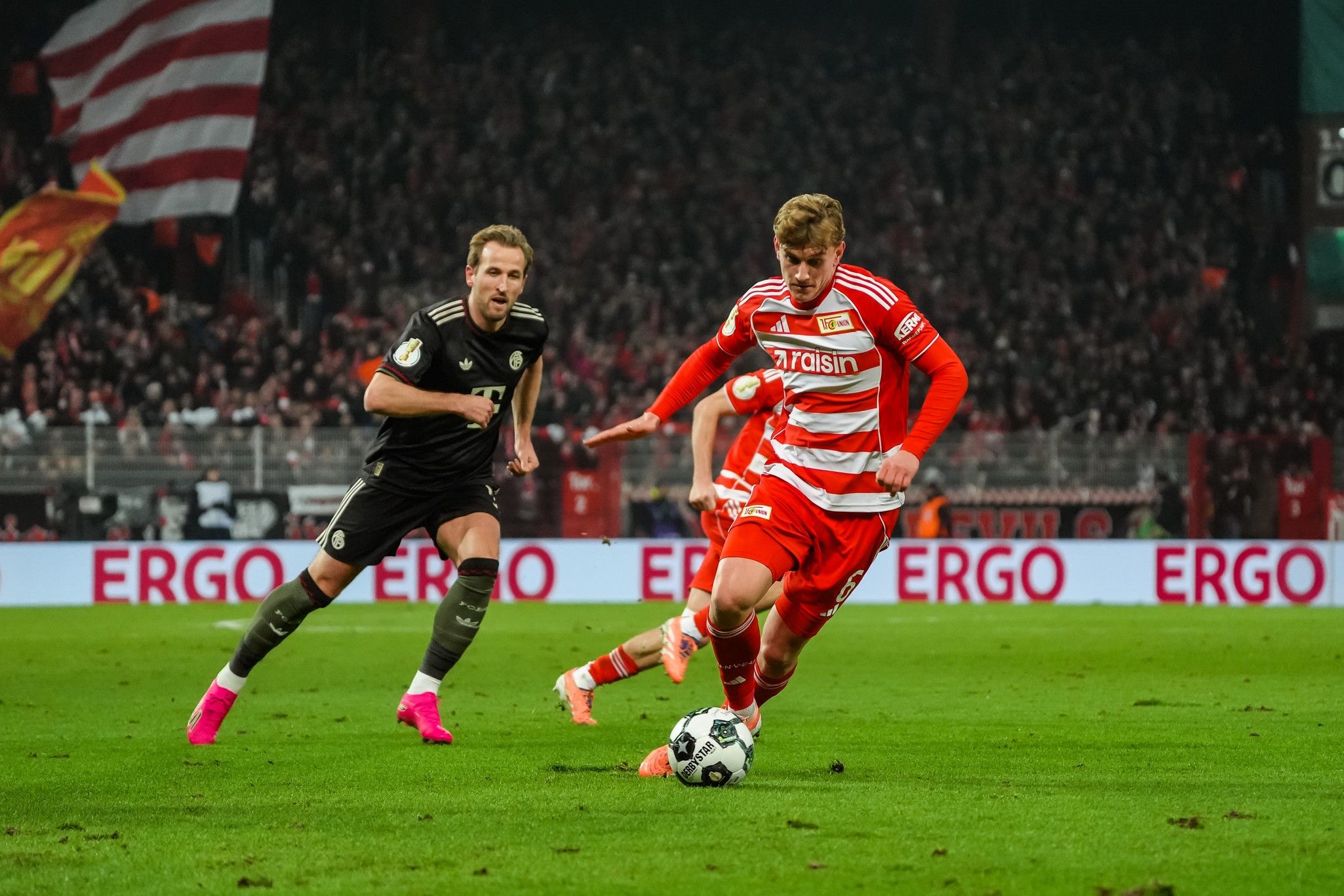 Spielszene im Fußball: Ein Spieler in rot-gestreiftem Trikot dribbelt den Ball, während ein Spieler in schwarz-gestreiftem Trikot verfolgt.