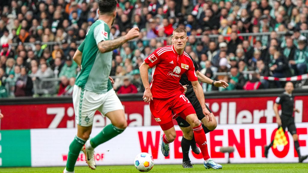Ein Fußballspieler in roter Uniform dribbelt den Ball auf einem Spielfeld mit Zuschauertribüne im Hintergrund.