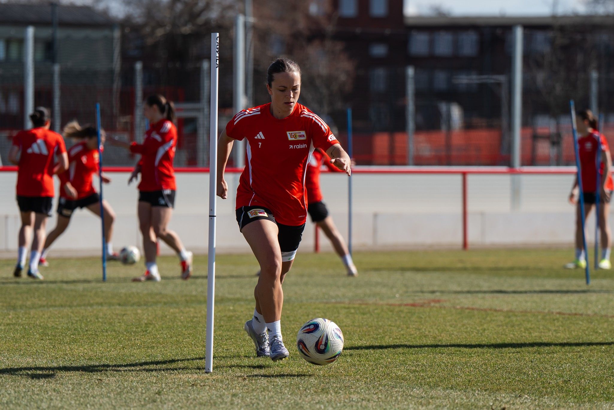 Spielerin in rotem Trikot dribbelt einen Fußball in einem Trainingsfeld, während andere Spieler im Hintergrund üben.