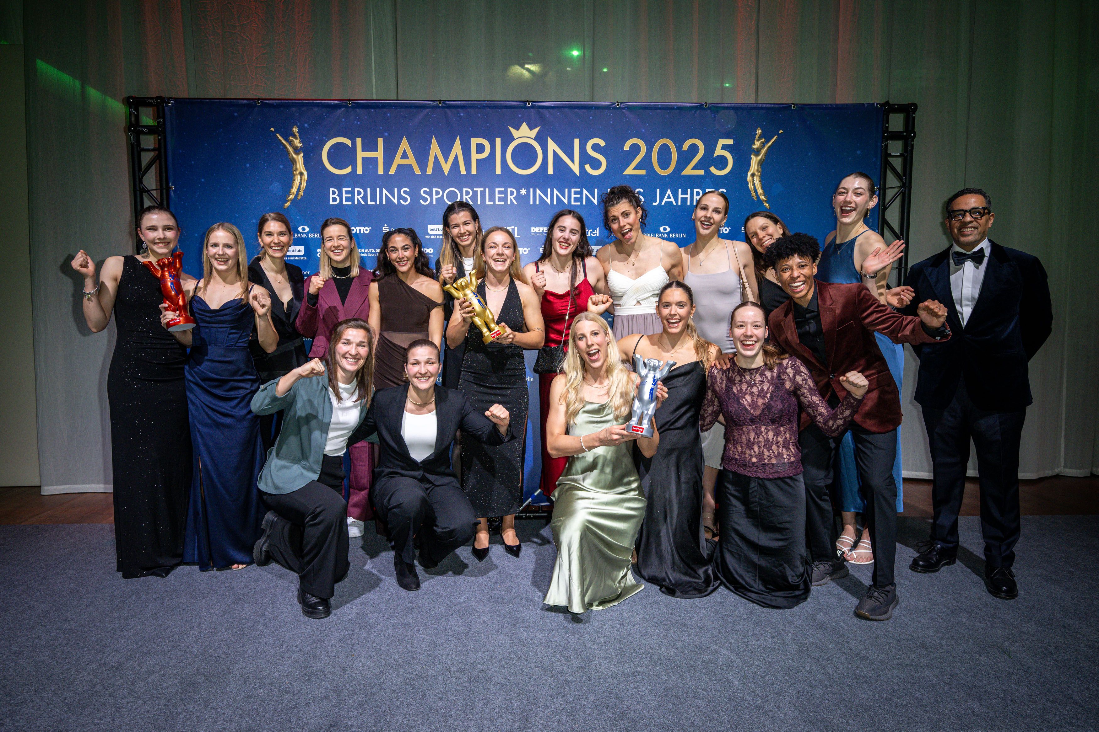 Gruppe von Menschen in festlicher Kleidung posiert lächelnd vor einer Hintergrundbühne mit dem Schriftzug „CHAMPIONS 2025“.
