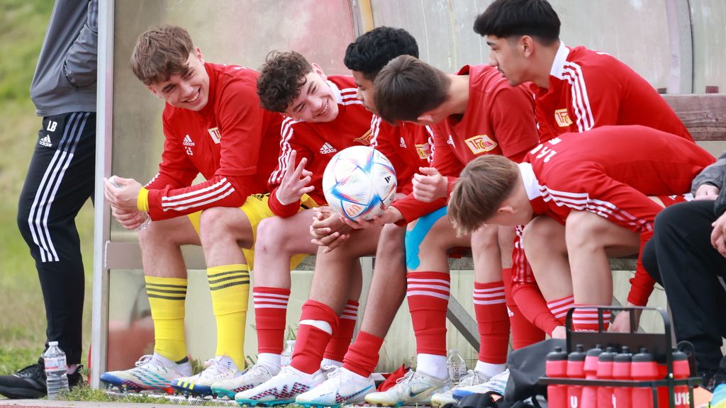 Sechs junge Fußballspieler in roten Trikots sitzen auf einer Bank, lachen und unterhalten sich, während einer einen Fußball hält.