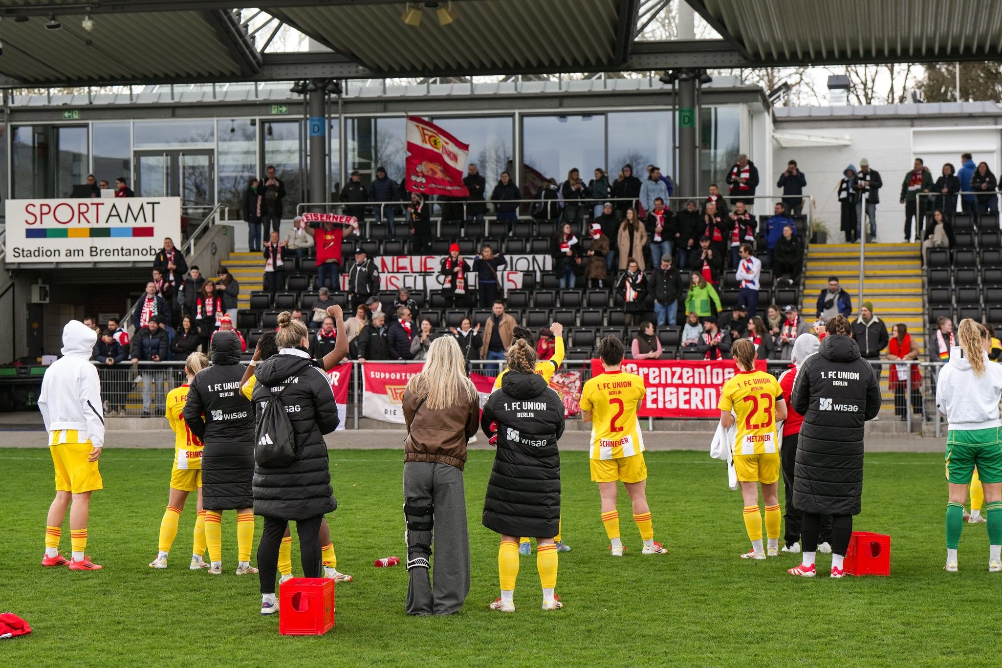 Eine Gruppe von Fußballspielern in gelben Trikots steht auf dem Spielfeld und verfolgt eine jubelnde Menge auf den Tribünen.
