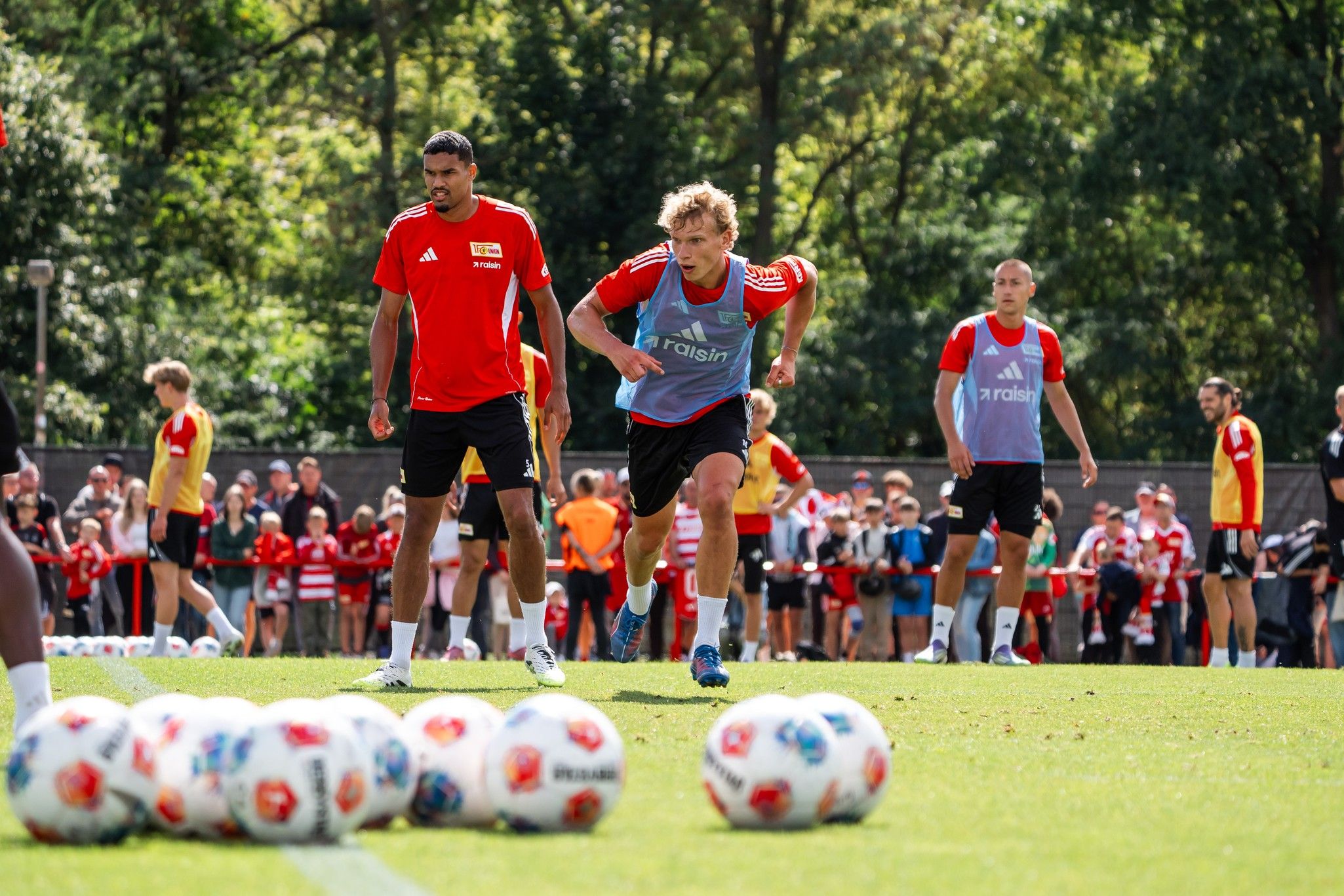 Spieler trainieren auf einem Fußballfeld, während Zuschauer im Hintergrund zuschauen. Bälle liegen in einer Reihe auf dem Rasen.