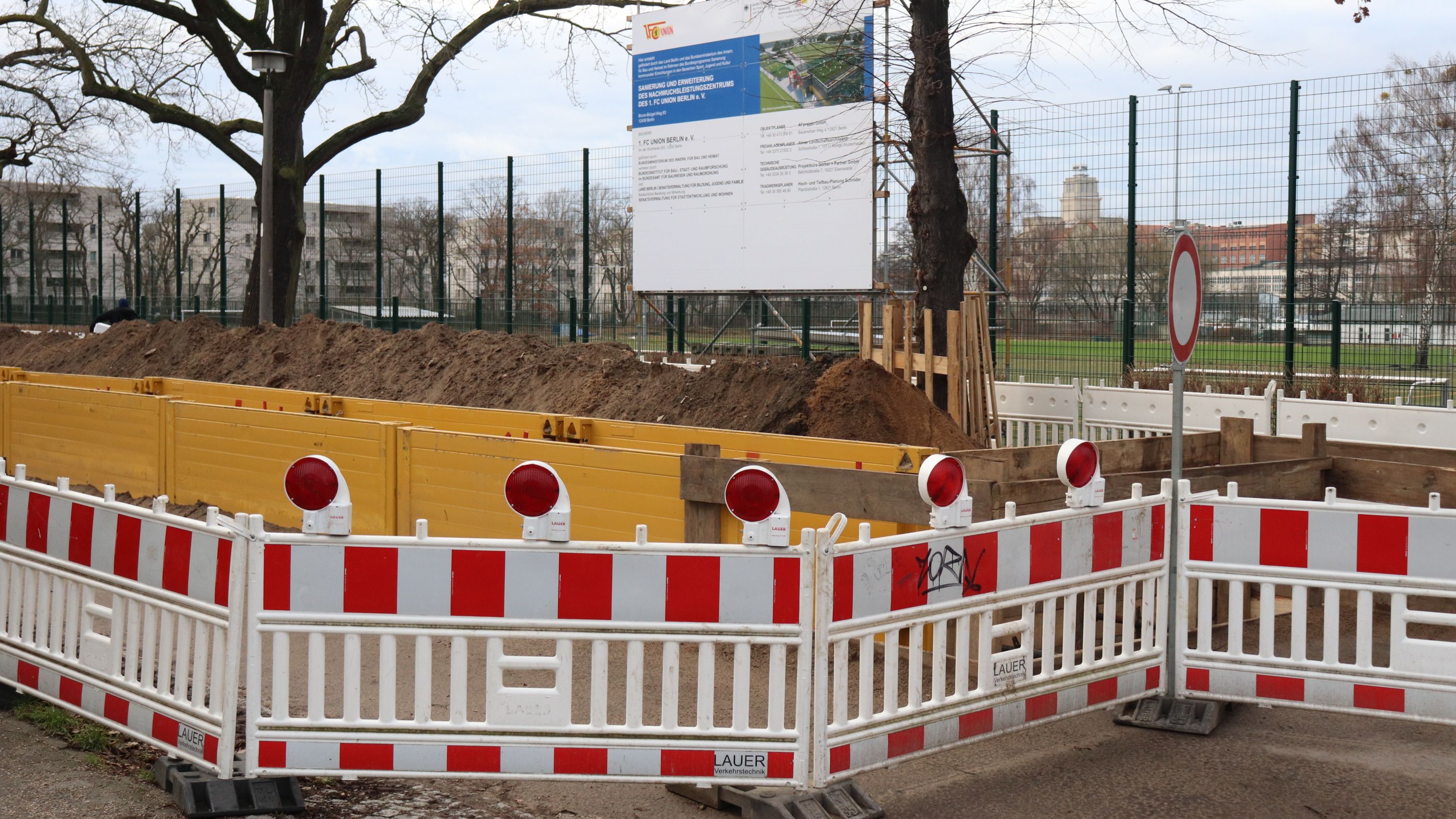 Baustelle mit Geländersperrung, einem großen Erdloch und einem Informationsschild im Hintergrund. Bäume und ein Sportplatz sind sichtbar.