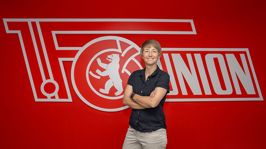 Person mit kurzem Haar steht vor einem roten Hintergrund mit dem Logo des FC Union Berlin.