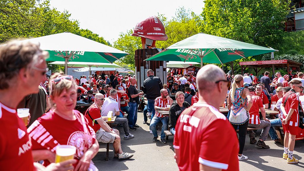 Menschenmenge in roten Trikots, die Getränke genießen und sich unter freiem Himmel auf einem Fest versammeln. Grüne Schirme spenden Schatten.