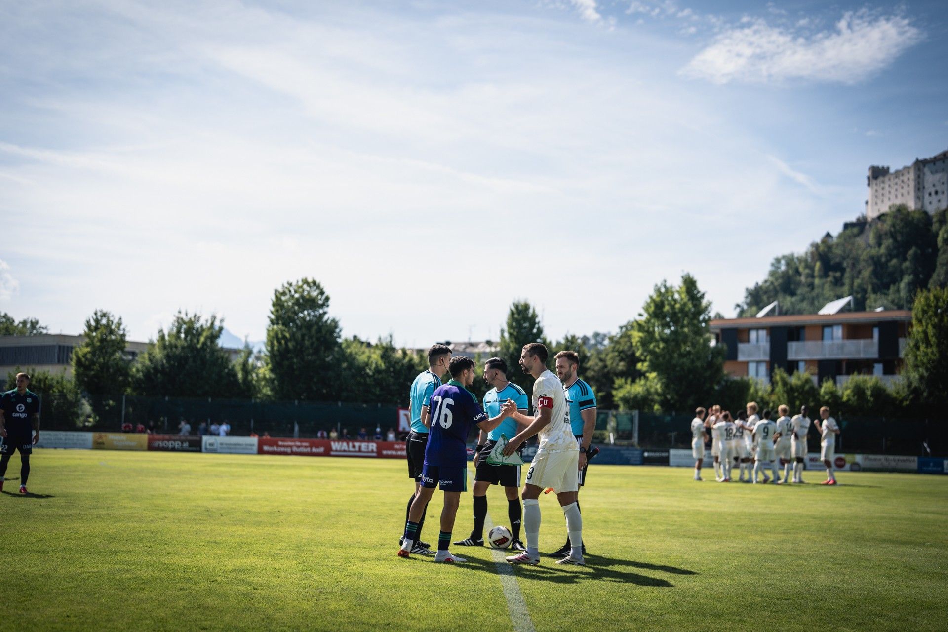 Zwei Gruppen von Fußballspielern schütteln sich vor dem Spiel die Hände auf einem sonnigen Rasenplatz. Im Hintergrund sind Zuschauer und Bäume zu sehen.