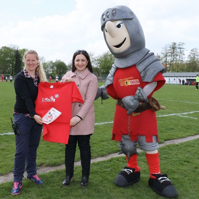 Frau in schwarzer Hose und rosa Jacke hält ein rotes T-Shirt, während ein Maskottchen und eine weitere Person daneben stehen.