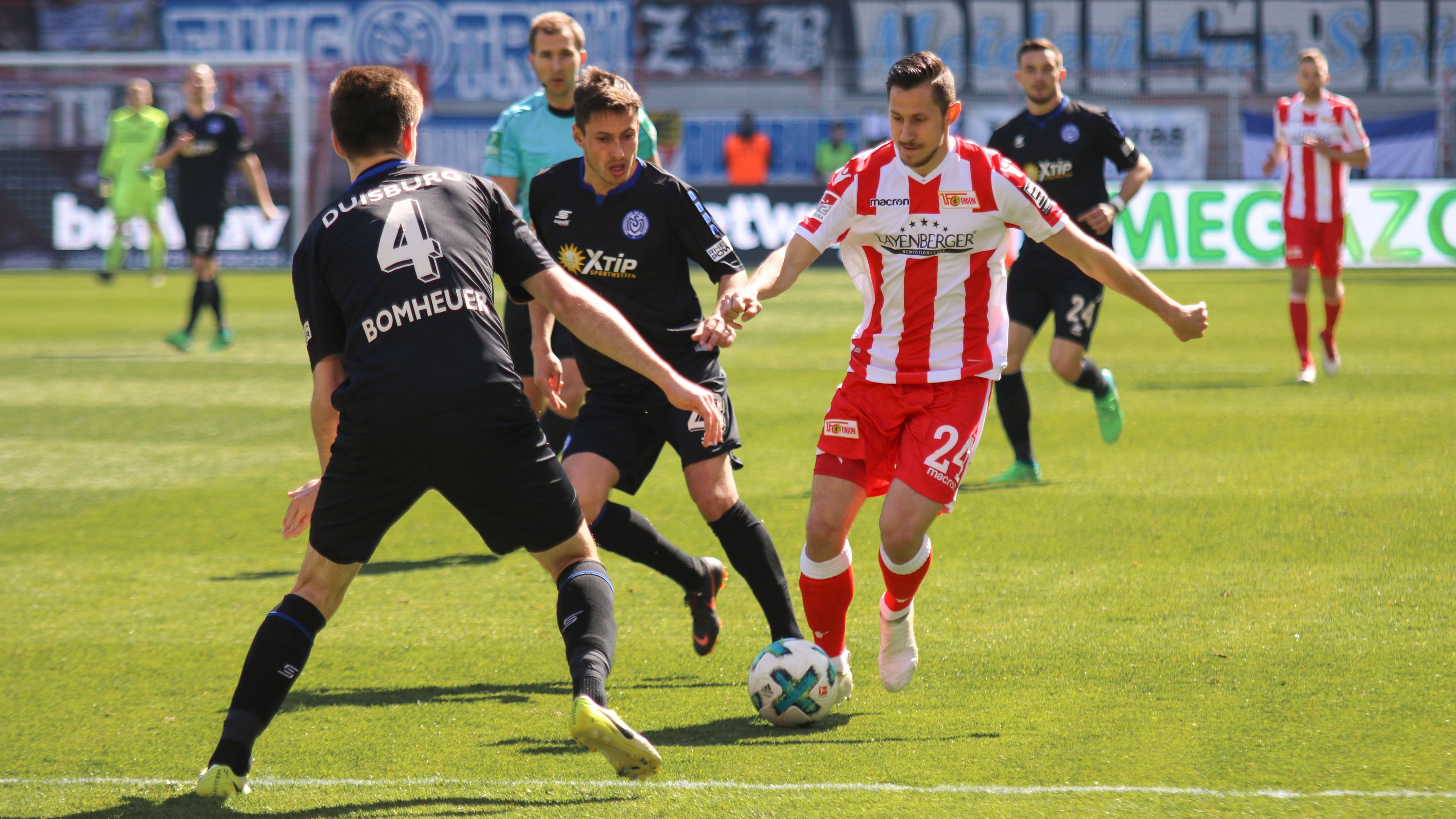 Fußballspiel mit zwei Spielern in Schwarzen und einem Spieler in Rot und Weiß, der den Ball dribbelt. Stadionankunft im Hintergrund.