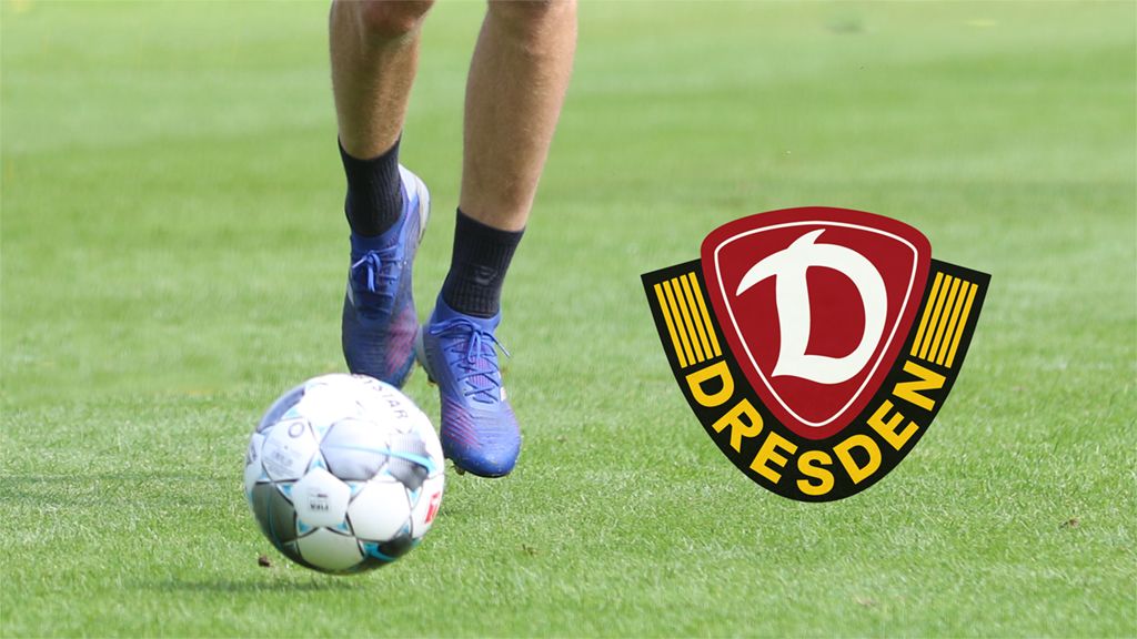 Players' feet dribble a soccer ball on green grass, with the Dynamo Dresden logo in the background.