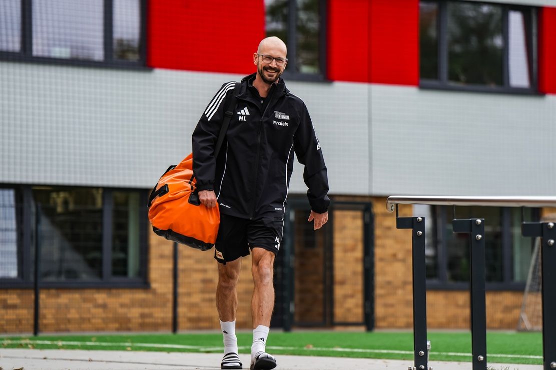 Ein Trainer mit Brille und Glatze trägt einen großen orangefarbenen Sportbeutel und geht lächelnd auf einem Gehweg entlang.