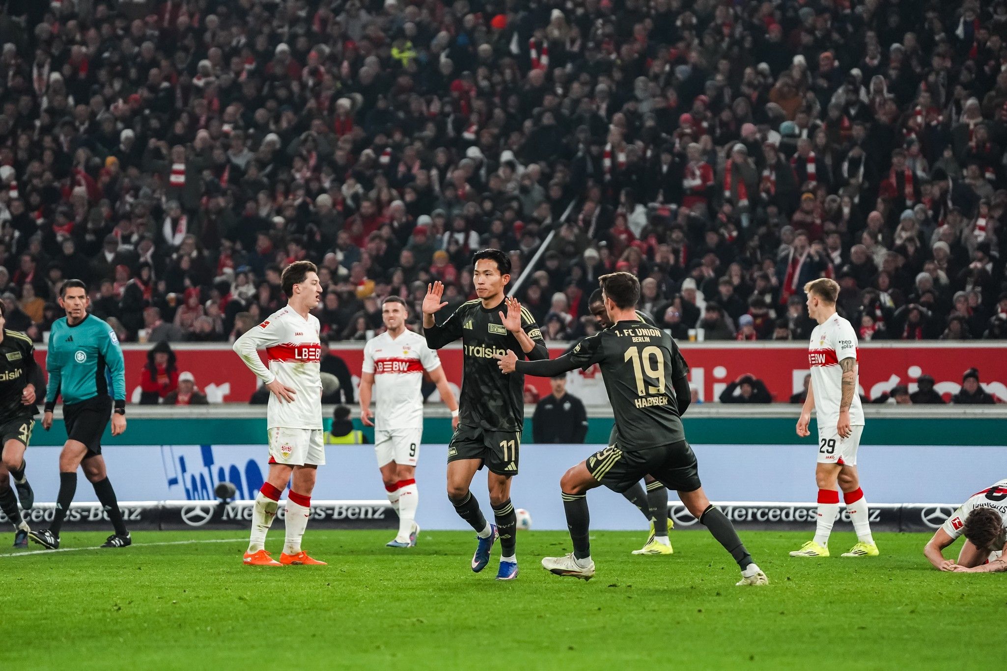 Spielszene mit Spielern in schwarzen und weißen Trikots; ein Torjubel und ein Spieler am Boden. Stadion voller Fans im Hintergrund.
