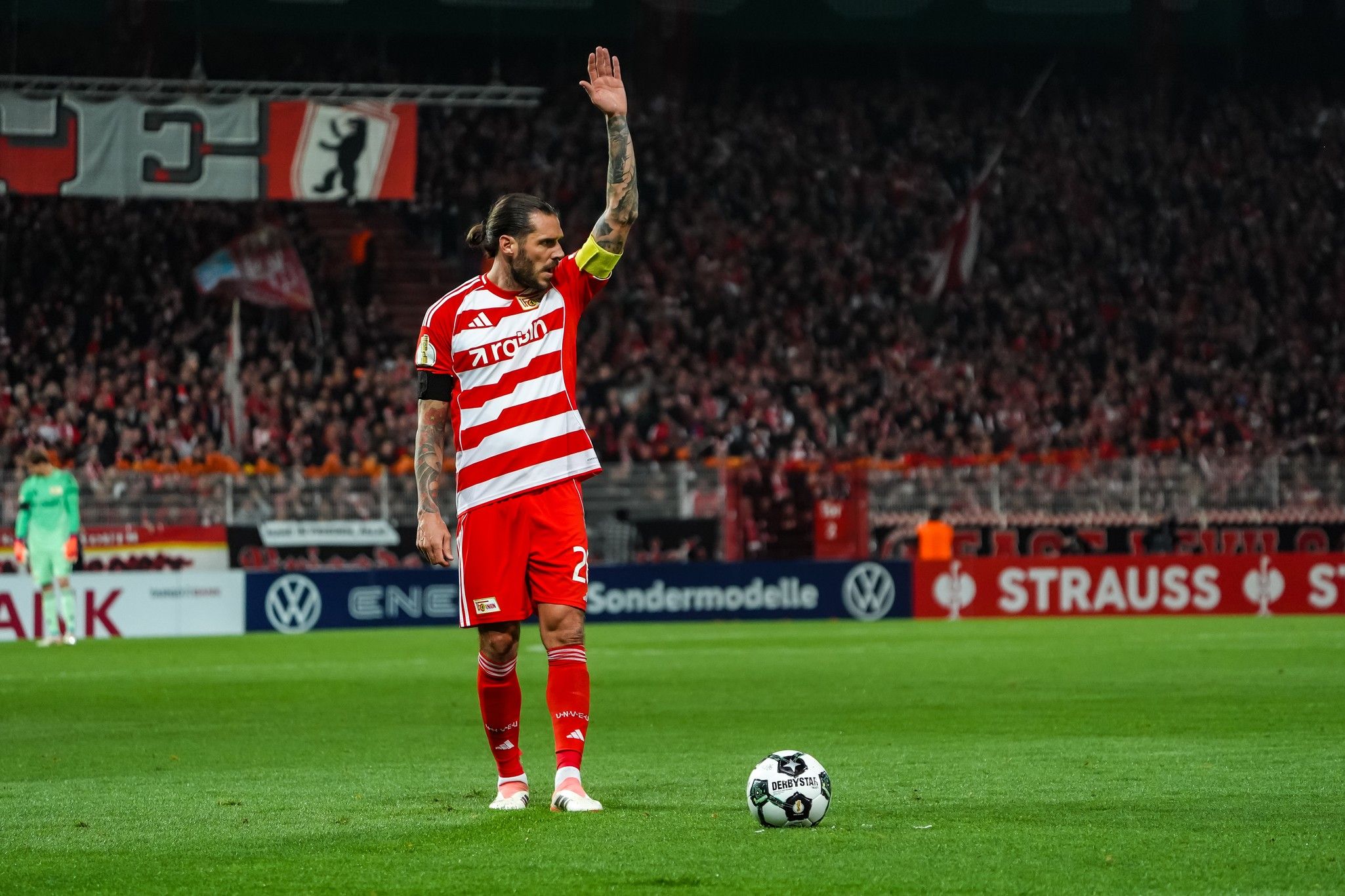 Ein Fußballspieler in rotem Trikot hebt die Hand, während er an der Seitenlinie steht, um das Publikum zu begrüßen.