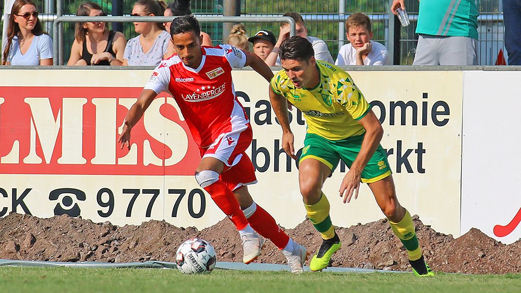 Two football players are fighting for the ball during a game, with spectators and an advertising banner in the background.