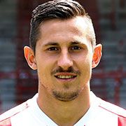 Eine Porträtaufnahme eines lächelnden Mannes mit kurzen, gestylten Haaren, der ein Fußballtrikot trägt.