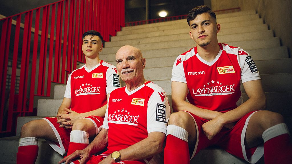 Drei Personen in Rot-Weiß, zwei junge Männer und ein älterer Mann, sitzen auf einer Treppe in Fußballtrikots.