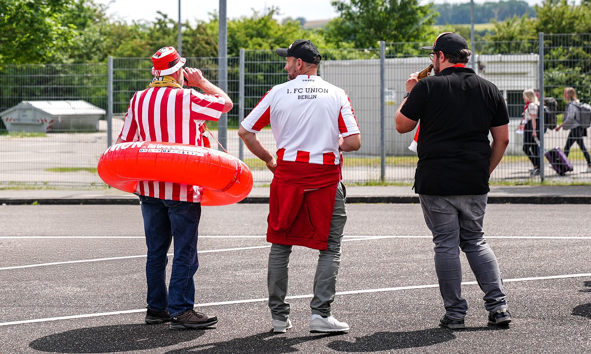 Drei Menschen stehen mit Blick auf ein Sportgelände, einer trägt einen Schwimmring, alle in Fußballtrikots.
