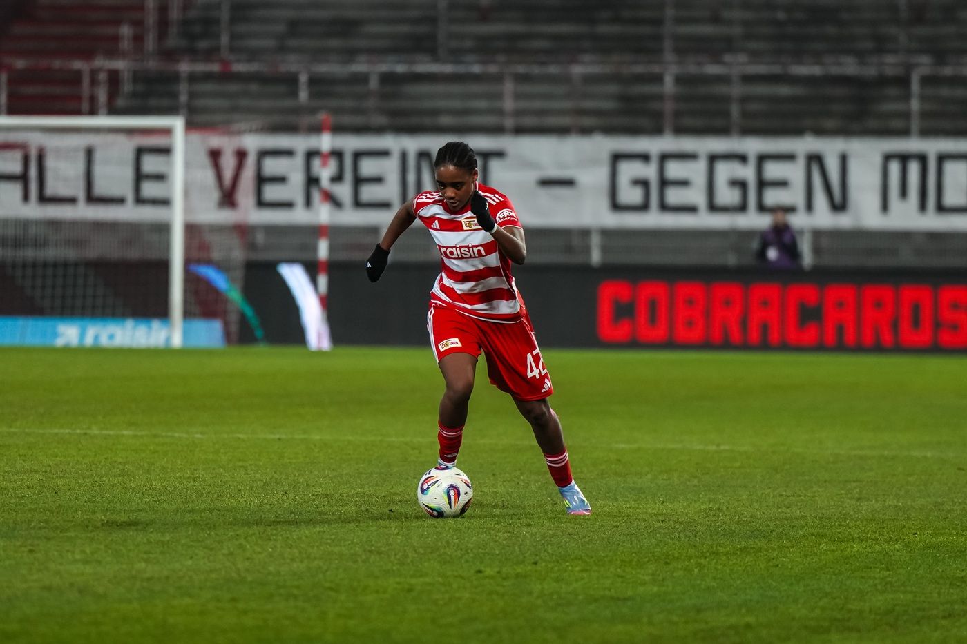 Eine Fußballspielerin in roter und weißer Uniform dribbelt den Ball auf einem Rasenplatz. Im Hintergrund ist ein Banner sichtbar.