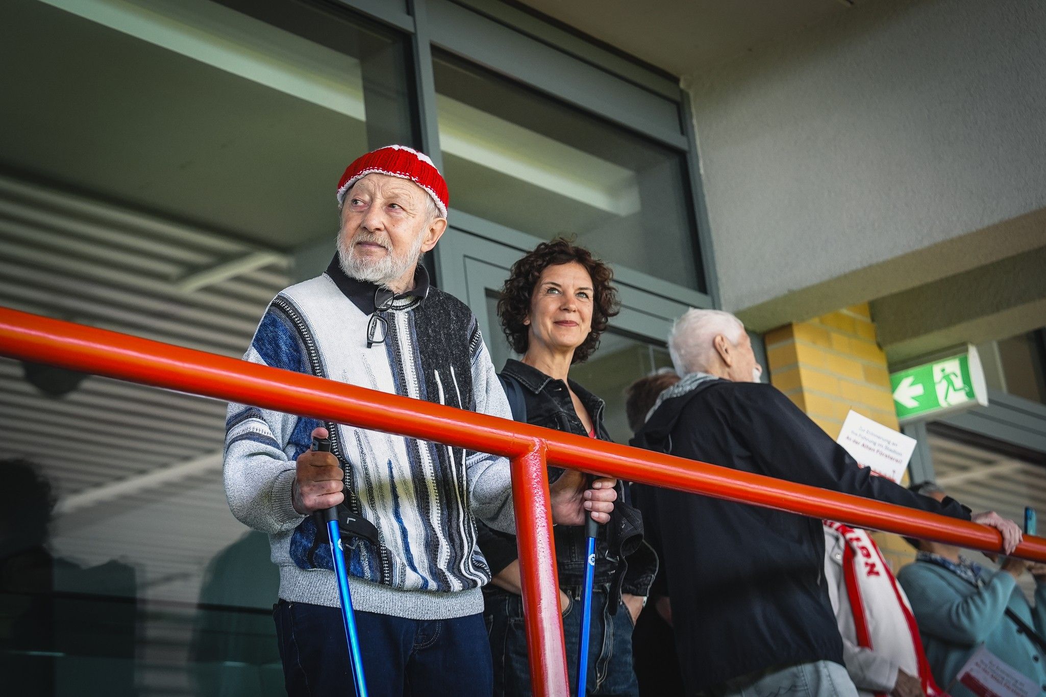 Ein älterer Mann mit rotem Hut und eine Frau stehen an einem Geländer in einem Stadion. Im Hintergrund sind weitere Zuschauer zu sehen.