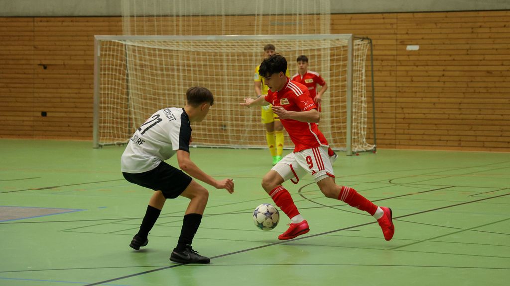 Zwei junge Fußballspieler in Hallenuniformen kämpfen um den Ball in einer Sporthalle. Im Hintergrund sind weitere Spieler sichtbar.