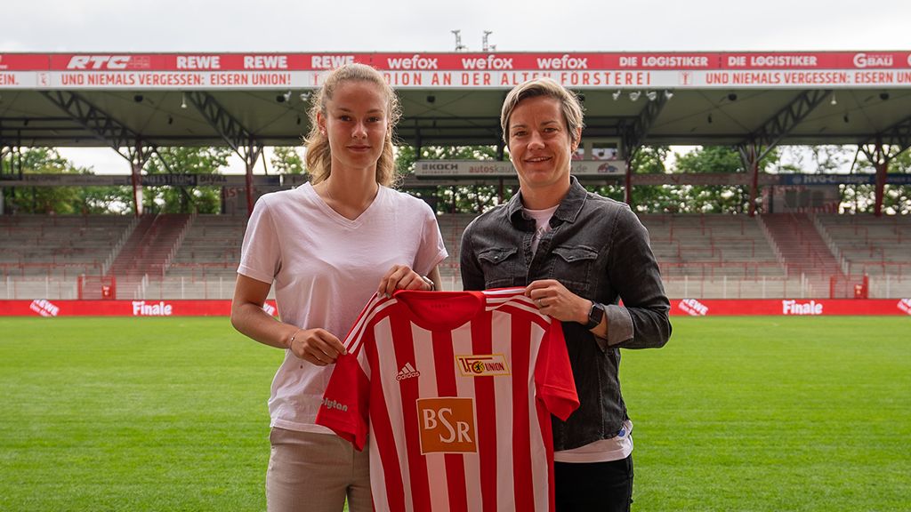 Zwei Frauen stehen auf einem Fußballfeld und halten ein Trikot mit roten und weißen Streifen in der Hand.