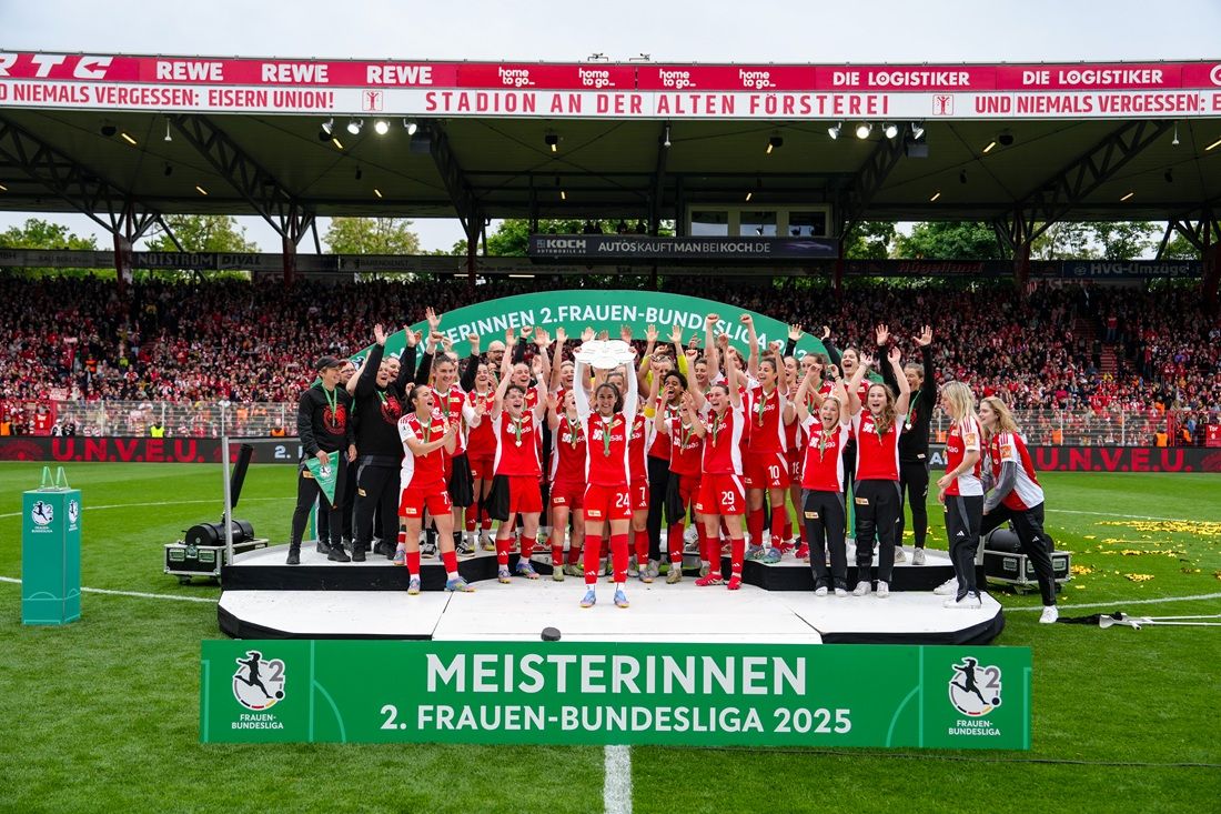 Fußballmannschaft feiert auf einer Bühne mit Pokal, umgeben von Fans und Konfetti bei der Meisterschaft der 2. Frauen-Bundesliga 2025.