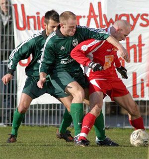 Zwei Spieler in grünen Trikots versuchen, einen Spieler in rotem Trikot am Ball zu hindern, während im Hintergrund ein Banner zu sehen ist.